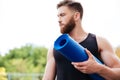 Serious male yoga instructor holding mat and looking away outdoors