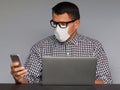 Man in eyeglasses and medical mask sitting at the desk in front of laptop and working online on his mobile phone at home. Royalty Free Stock Photo