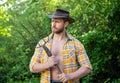 Serious man holding hayfork. Unshaven man wearing cowboy hat and plaid shirt outdoors. Man farmer