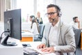 Serious man wearing formal clothes and headset looking at computer screen in bright office. Royalty Free Stock Photo