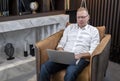 Serious mature businessman sitting in comfortable armchair and keeping laptop on lap. Man working or studying at home or Royalty Free Stock Photo