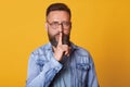 Serious middle aged emotionless bearded man in blue shirt, showing silence gesture, asks to keep secret, putting forefinger on Royalty Free Stock Photo