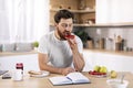 Serious millennial caucasian guy with beard eats sandwich with jam, reads book at table in kitchen interior