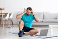 Serious millennial european male athlete puts headphones sits on mat in living room interior with bottle