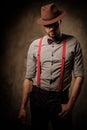 Serious old-fashioned man with hat wearing suspenders and bow tie, posing on dark background.