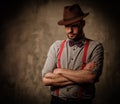 Serious old-fashioned man with hat wearing suspenders and bow tie, posing on dark background.