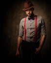 Serious old-fashioned man with hat wearing suspenders and bow tie, posing on dark background.