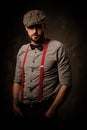 Serious old-fashioned man in tweed hat wearing suspenders and bow tie, posing on dark background.