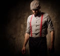 Serious old-fashioned man in tweed hat wearing suspenders and bow tie, posing on dark background.