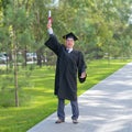 A serious old man in a graduate robe pulls his hand with a diploma up outdoors.