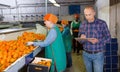 Serious owner of fruit warehouse checking work of female employees engaged in tangerines sorting