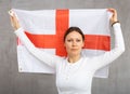 Serious patriotic adult woman holding state flag of England against gray wall background indoors