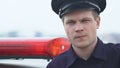 Serious patrolman in hat standing near squad car and looking to camera, enforcer