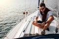 Serious and peaceful guy sits on board of yacht. He holds and looks at tablet. Young man is calm. He wears white sirt Royalty Free Stock Photo