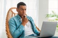 Serious pensive handsome young black man in casual looks at laptop, sits in chair in bright room interior Royalty Free Stock Photo