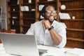 Serious Positive and cheerful african-american businessman in headset sitting in front of the laptop and talking online Royalty Free Stock Photo