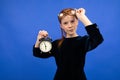 Serious red-haired teenager girl in glasses in a black dress recalls punctuality holding an alarm clock on a blue