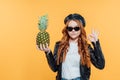 Serious redhead girl in leather jacket holding pineapple and showing ok sign isolated on yellow.