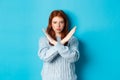 Serious redhead girl looking confident, showing cross gesture to stop and forbid action, standing over blue background