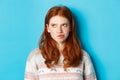 Serious redhead girl thinking, grimacing and frowning while pondering, looking left at copy space, standing over blue