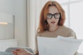 Serious redhead young woman focused in paper documents studies financial report