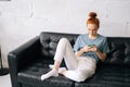 Serious redhead young woman is typing message on cell phone while sitting on soft black couch Royalty Free Stock Photo