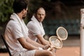 serious retro styled tennis players sitting on chairs with towels and rackets at tennis