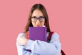 Serious school girl with blond hair and intent look keeping many books in her hands with backpack and eyeglasses.