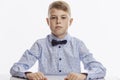 A serious schoolboy in a blue shirt with a bow tie sits at the table. White background Royalty Free Stock Photo
