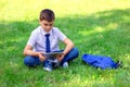 Serious Schoolboy in a white shirt and blue tie sits on green grass and plays with a tablet Royalty Free Stock Photo