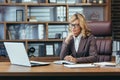 Serious senior business woman in a suit sitting in the office at a desk with a laptop and making notes in a notebook Royalty Free Stock Photo