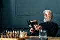 Serious senior gray-haired older male reading paper book at home sitting at wooden table with chess board, relax and Royalty Free Stock Photo