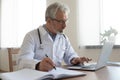 Serious senior physician using computer app making notes in journal