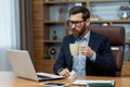 Serious and shocked young man working in the office, sitting at the desk, holding a cup with a drink in his hand and Royalty Free Stock Photo
