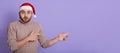 Serious and shocked young Santa man in beige sweater and Christmas hat, pointing index fingers aside to copy space for promo,