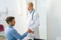 Serious standing mature male adult doctor examining wrist of suffering male patient sitting at table during checkup