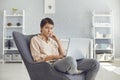 Serious woman looking at camera while working on laptop sitting in cozy armchair