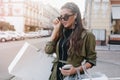 Serious tanned woman in dark glasses listening music in white earphones. Charming hispanic lady in trendy jacket