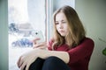 A serious teenager girl with long blond hair is sitting by the window on a windowsill with a phone in her hand. The concept of