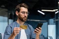 Serious thinking man inside office holding bank credit card and smartphone, businessman making money transfer Royalty Free Stock Photo