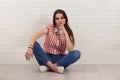 Serious thinking young woman sitting cross-legged on floor, touching chin, looking at camera, isolated white brick wall background Royalty Free Stock Photo