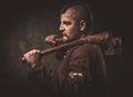 Serious viking with ax in a traditional warrior clothes, posing on a dark background.