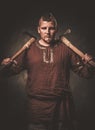 Serious viking with axes in a traditional warrior clothes, posing on a dark background.