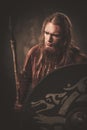 Serious viking with a spear in a traditional warrior clothes, posing on a dark background.