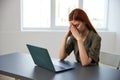 a serious, work-weary woman, with a sad face, sits at a laptop with her hands folded near her face