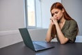 a serious, work-weary woman, with a sad face, sits at a laptop with her hands folded near her face