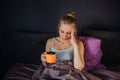 Serious and worried young woman sits on bed. She holds cup of tea with lemon and holds hand on forehead. She has