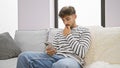 Serious young arab man doubting his next idea, deeply immersed while texting on his smartphone, sitting solo on the living room