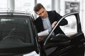 Serious young auto insurance agent checking new car, taking notes, working at automobile dealership