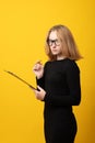 Serious young blond woman in black dress, glasses. Writing notes in notebook, looking aside